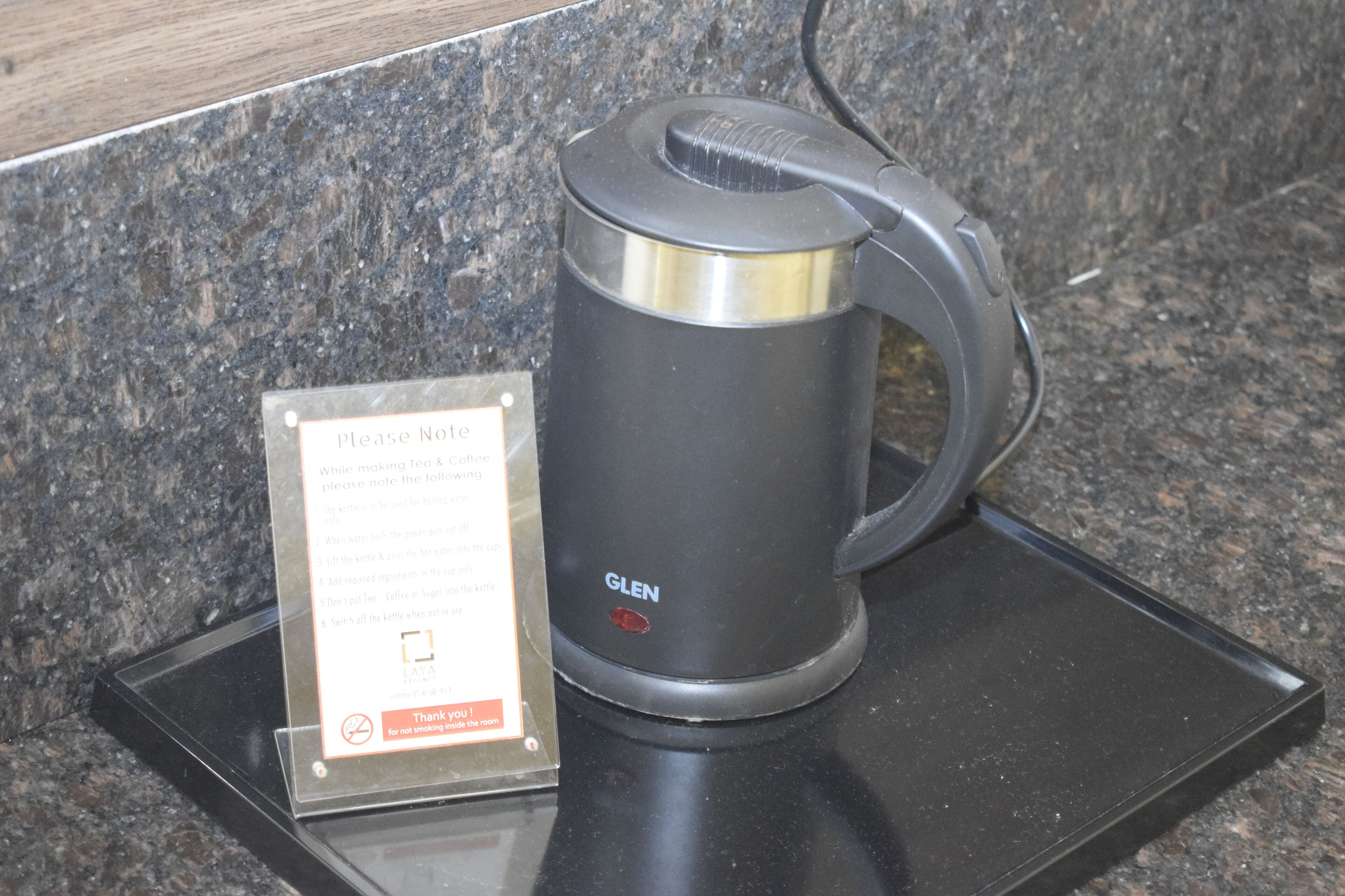 A black GLEN electric kettle on a granite countertop with a sign instructing not to smoke and to note the kettle's location.
