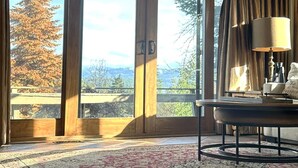 A room with large sliding glass doors, a patterned rug, a table with a lamp, and a view of trees and mountains.