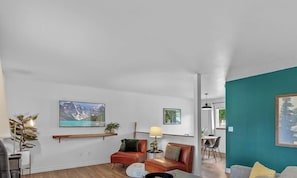 A living room with a sofa, a coffee table, a TV mounted on the wall, and a dining area visible through an open door.