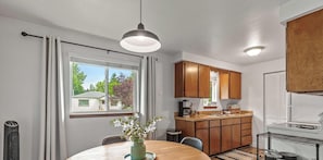 A modern kitchen with wooden cabinets, a dining area with a table and chairs, and a large window with a view of houses.