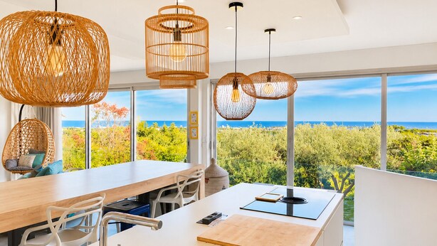 A modern kitchen with a large island, hanging wicker light fixtures, and a view of the ocean.