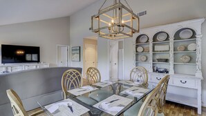 A dining area with a glass table, wooden chairs, a hanging light fixture, and a white cabinet with decorative plates.