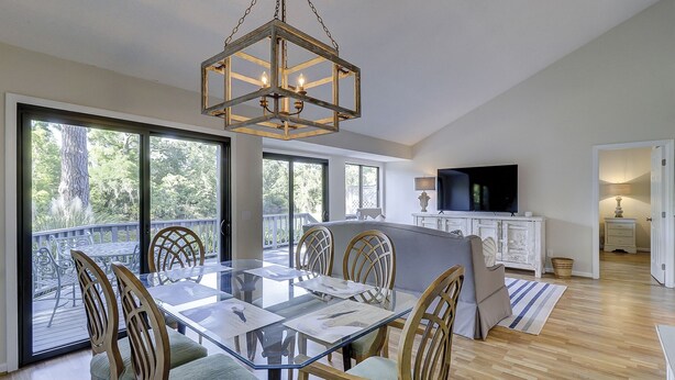 A spacious dining area with a glass table and wooden chairs, a large TV on a white cabinet, and a balcony with a view of trees.
