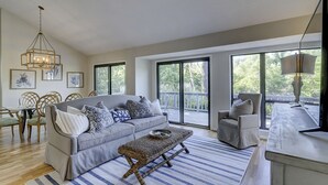 A spacious living room with a grey sofa, wicker coffee table, and a dining area with wooden chairs. Large windows offer views of the outdoors.
