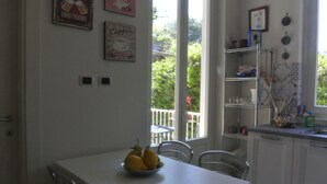 Een keuken met een witte tafel, stoelen en een schaal met citroenen op de tafel. Er zitten twee lichtschakelaars op de muur en een raam met uitzicht op groen buiten.