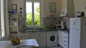 Een kleine keuken met een wasmachine, een magnetron en een koelkast. Er is een raam dat natuurlijk licht en uitzicht op groen biedt.