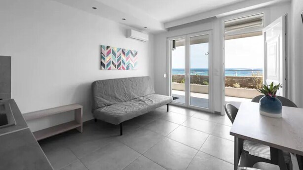 Un salon moderne avec un canapé gris, une table à manger et des chaises, une œuvre d’art colorée accrochée au mur et une grande porte-fenêtre coulissante donnant sur un balcon avec vue sur la mer.