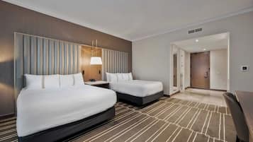 A hotel room with two beds, a bedside table with a lamp, a carpeted floor, and a door leading to another room.