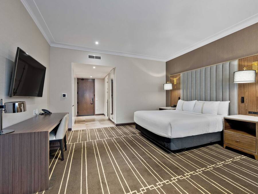 A hotel room with a bed, bedside table, lamp, desk with chair, and a TV mounted on the wall.
