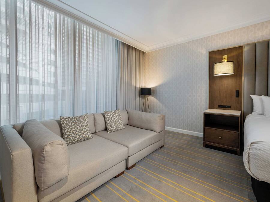 A hotel room with a sofa, a bed, a nightstand, a lamp, and a window with curtains.