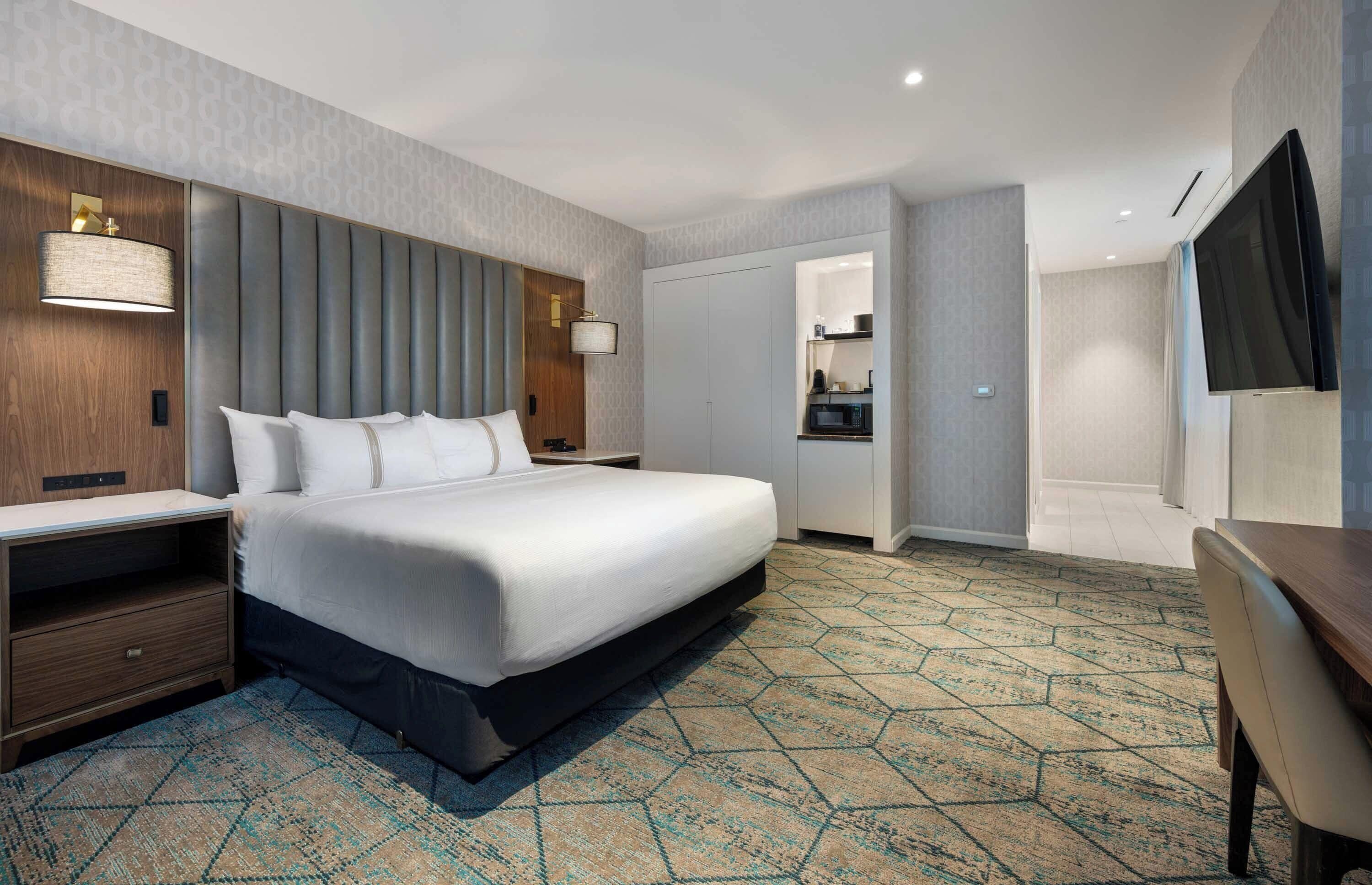 Habitación de hotel moderna con una cama grande, un escritorio, un televisor de pantalla plana y una kitchenette al fondo.