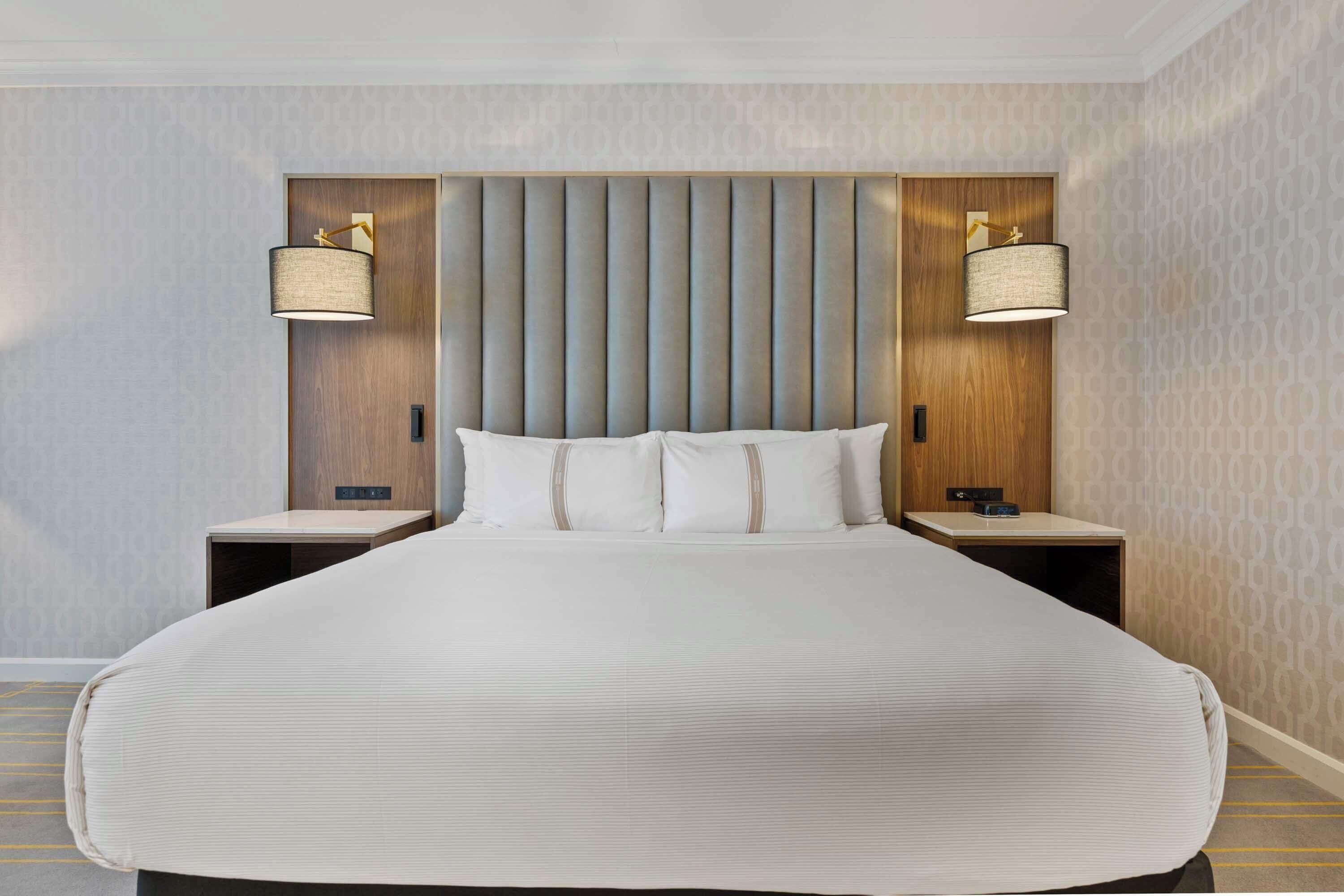 Habitación de hotel con una cama grande, dos mesitas de noche, cabecero con listones verticales y dos lámparas de pared.