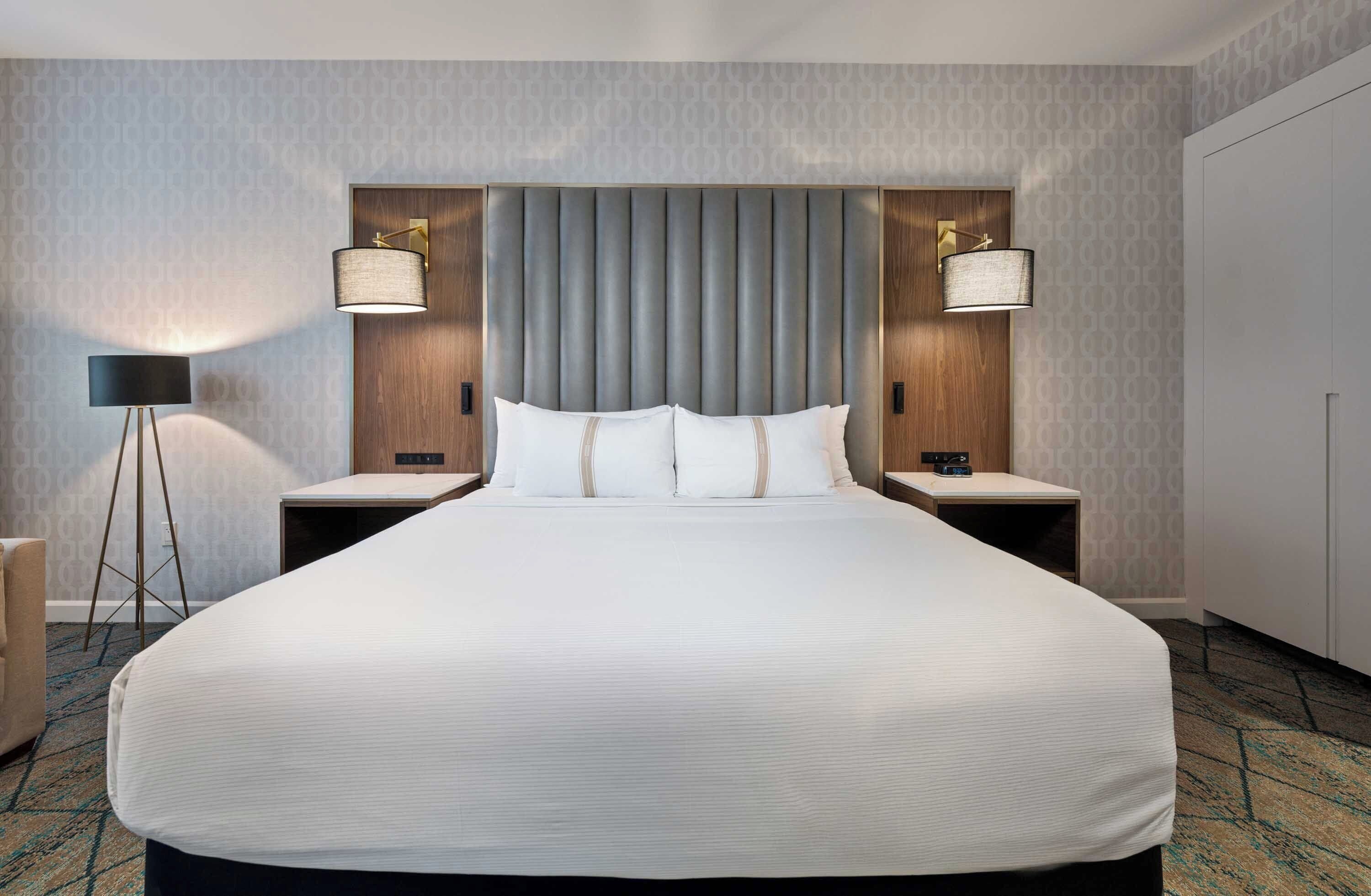A large bed with a grey headboard, two bedside tables, two lamps, and a carpeted floor.