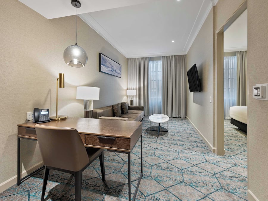 A hotel room with a desk, chair, two lamps, a sofa, a small round table, and a TV mounted on the wall.