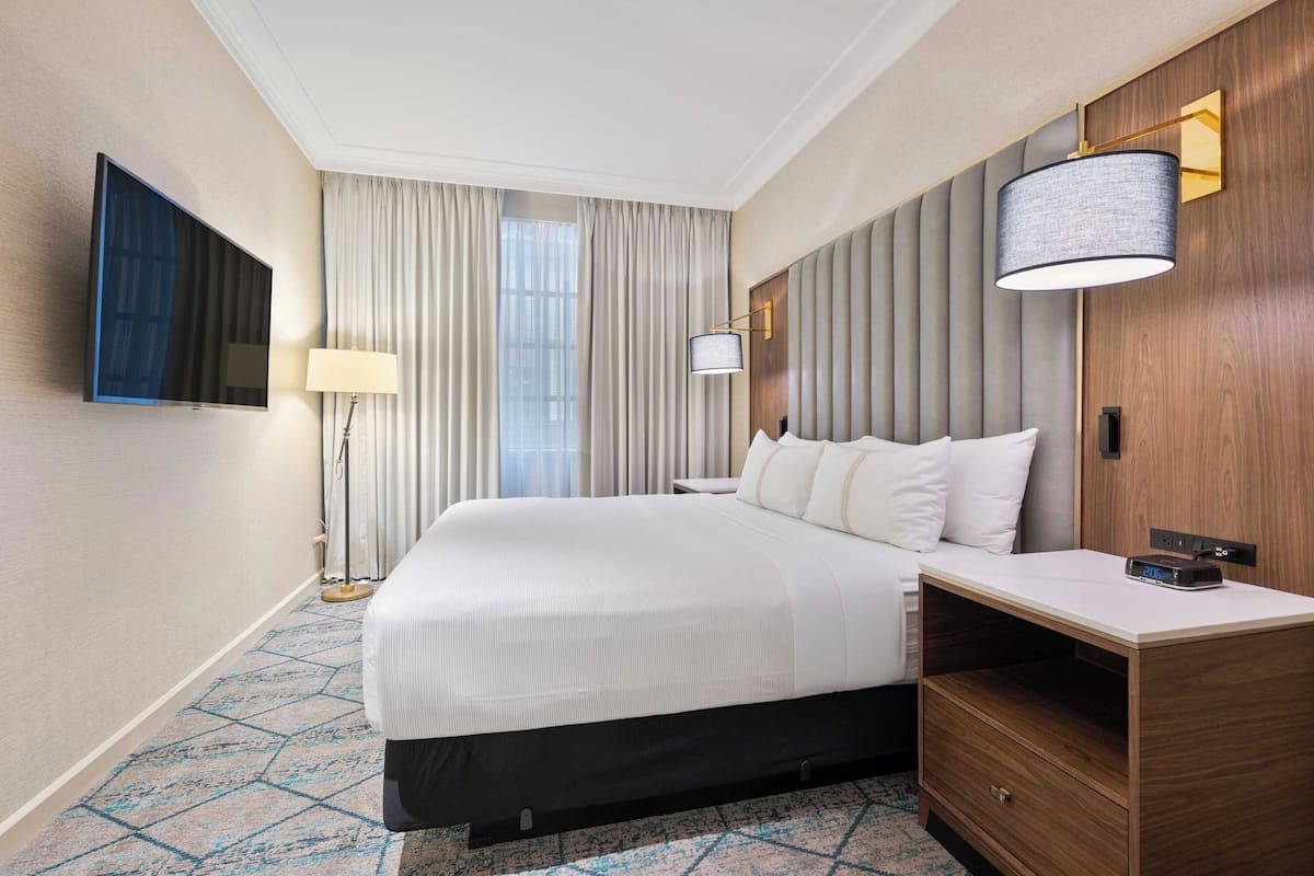 Habitación de hotel con una cama grande, un televisor de pantalla plana montado en la pared, una mesita de noche con una lámpara y un cabecero de madera.