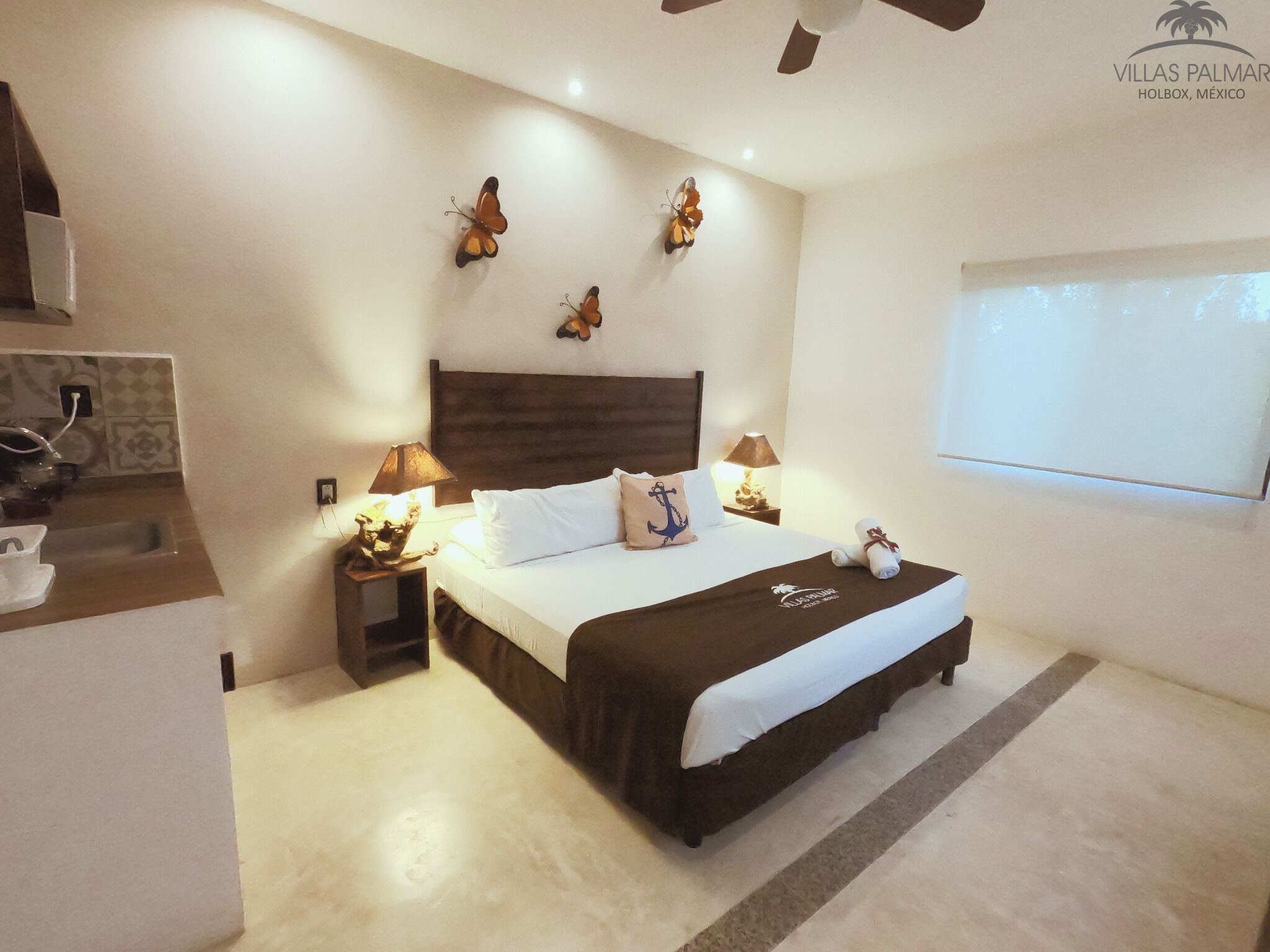 A hotel room with a bed, two bedside tables, a ceiling fan, decorative butterfly wall art, and a window with a view.