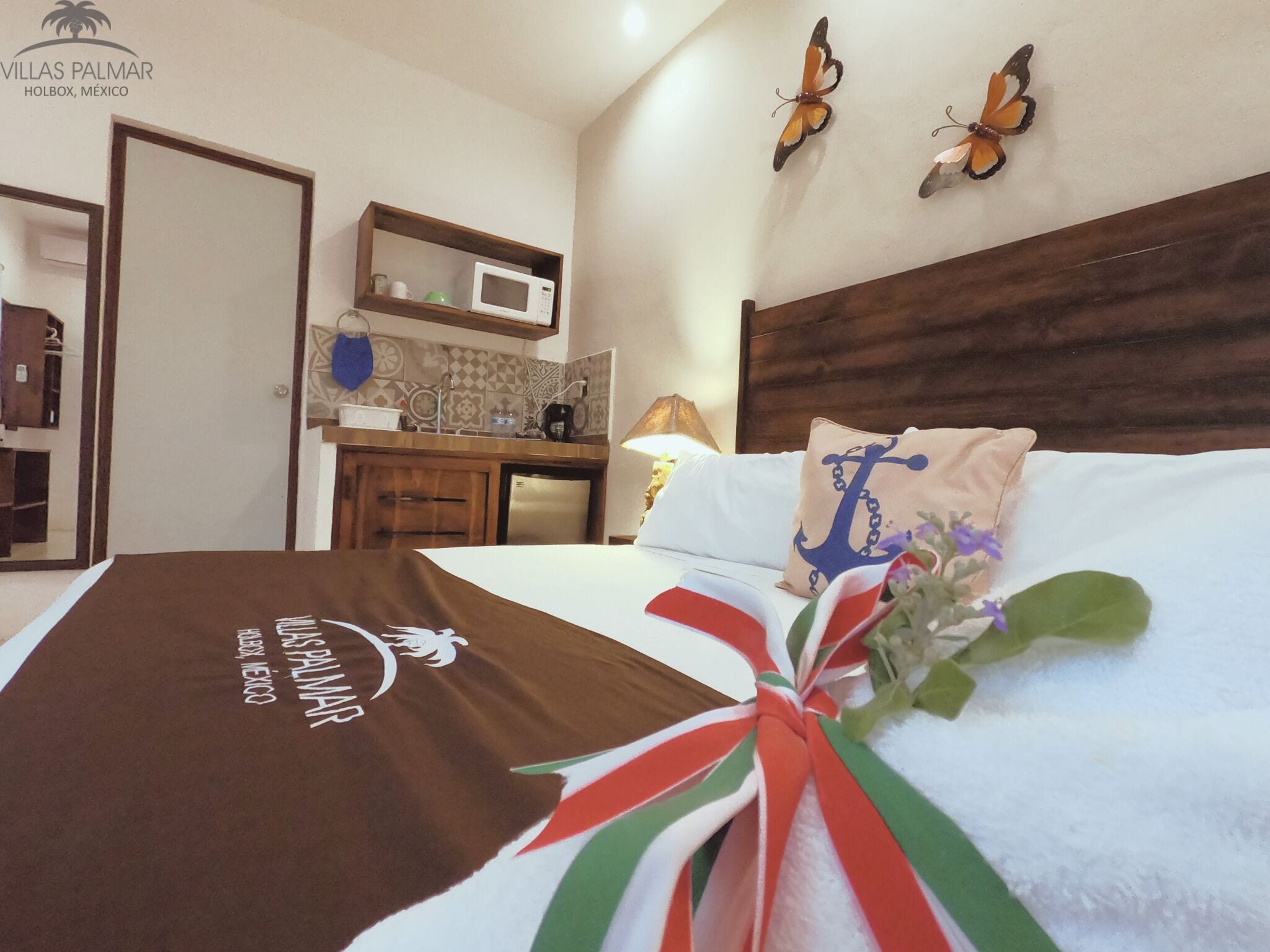 Habitación de hotel con cama, almohada con diseño de ancla, cojín decorativo con estampado de bandera, decoración de mariposa en la pared y una kitchenette.