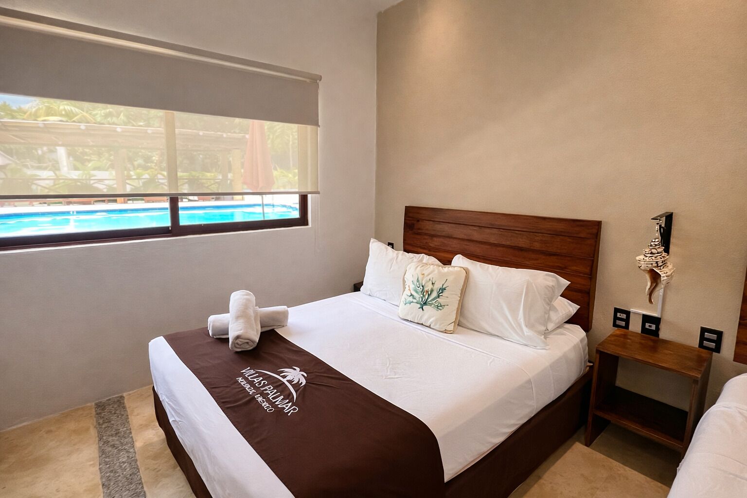 A hotel room with a bed, a nightstand, a window with a view of a pool, and a decorative wall piece.