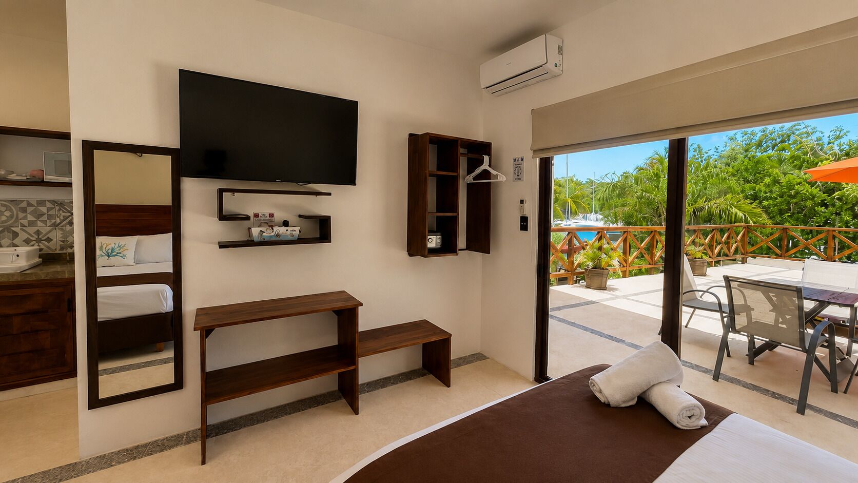 Une chambre d’hôtel comprenant un lit, une télévision, un banc en bois, un miroir, une étagère avec des articles de toilette et une vue sur un patio extérieur avec une table et des chaises.