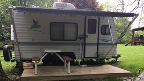 Ein Wohnmobil mit einer kleinen Terrasse und Tisch, umgeben von Gras und Bäumen.
