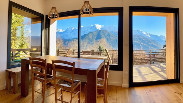 A dining area with a wooden table and chairs, large windows with mountain views, and a hanging light fixture. - 🏔 Symphonie des Cimes - fully renovated 2024 | Rando & Tennis  (Passy)