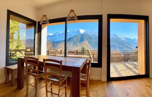 A dining area with a wooden table and chairs, large windows with mountain views, and a hanging light fixture. - 🏔 Symphonie des Cimes - fully renovated 2024 | Rando & Tennis  (Passy)