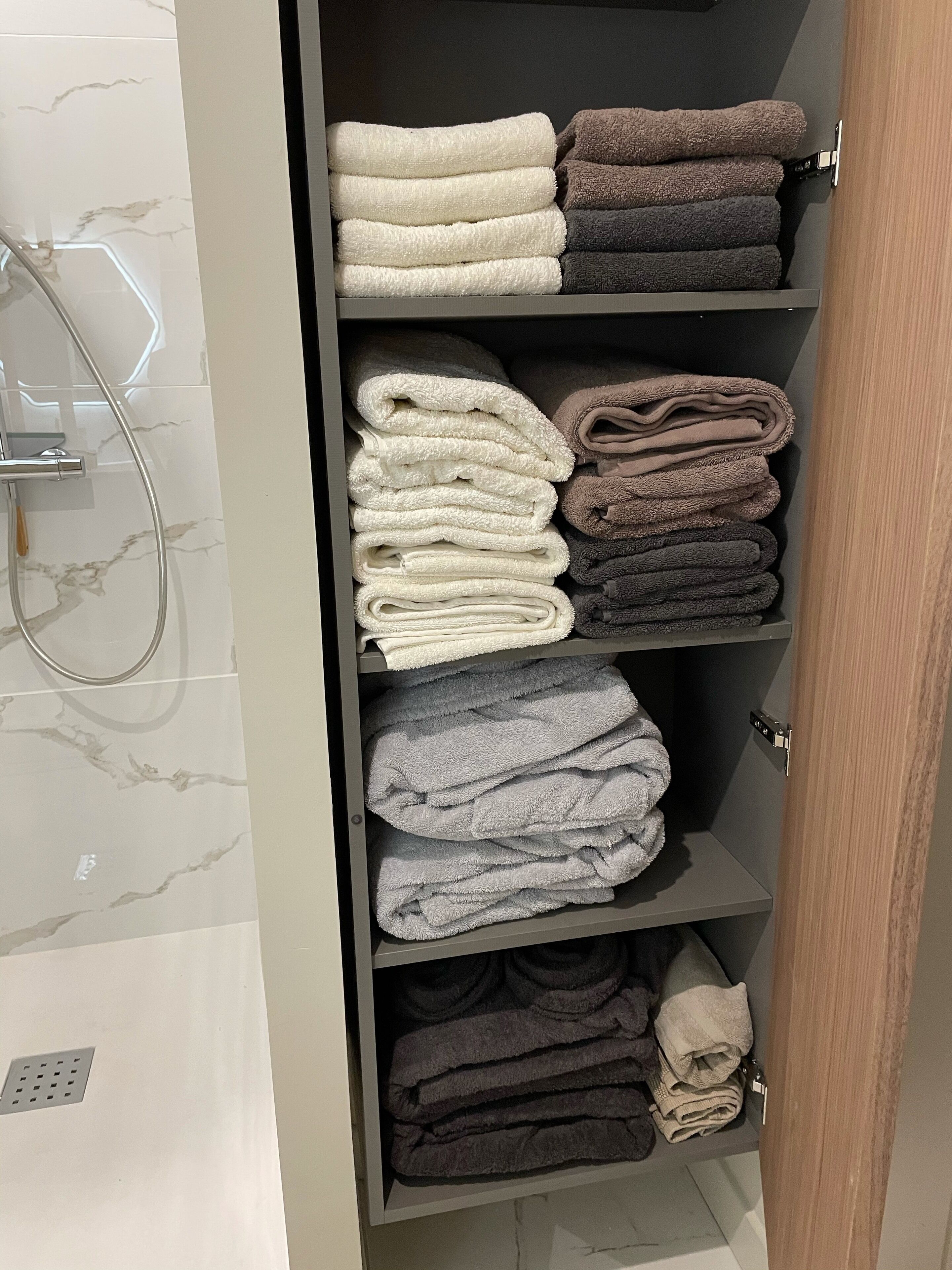 Une salle de bain avec un mur recouvert de carreaux de marbre, une douche avec une porte en verre et une étagère où sont pliées avec soin des serviettes de différentes couleurs.