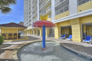 A hotel pool area with a water fountain, lounge chairs, and a building with balconies.