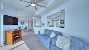 A spacious living room with a large blue sofa, a wooden entertainment unit, a ceiling fan, and a large mirror. - Horizon Penthouse 18, Lovely Condo in Myrtle Beach (Myrtle Beach)