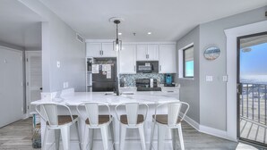 A modern kitchen with white cabinetry, a large island with bar stools, a microwave, and a view of the ocean through a large window. - MB Resort Renaissance 514 - OV 2 Bedrooms (Myrtle Beach)