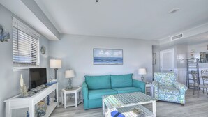 A living room with a teal sofa, a coffee table, a TV, and a painting of the ocean. - MB Resort Renaissance 514 - OV 2 Bedrooms (Myrtle Beach)
