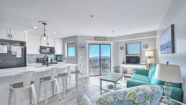 A modern kitchen with white cabinets, a microwave, and a dining area with white chairs. There's a teal sofa and a lamp in the living area. - MB Resort Renaissance 514 - OV 2 Bedrooms (Myrtle Beach)
