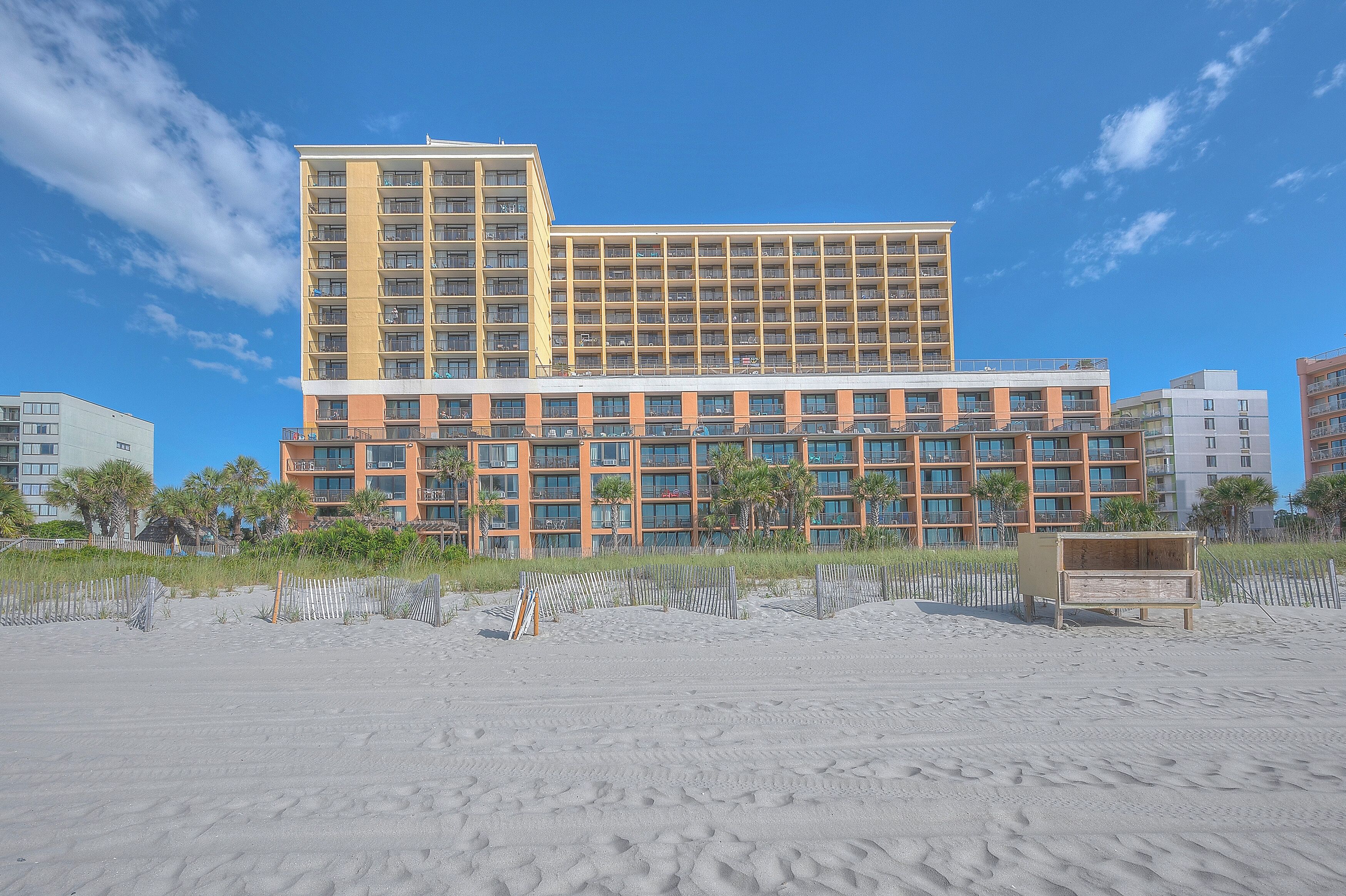 Ein Hotel mit mehreren Etagen, einem Sandstrand davor und einem strahlend blauen Himmel.