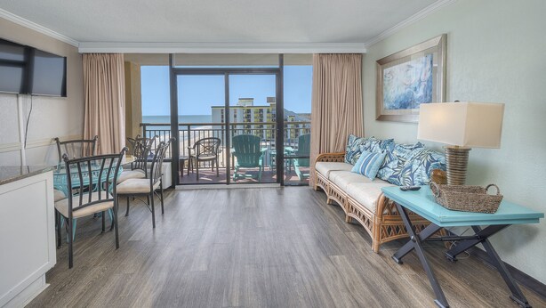 Ein Hotelzimmer mit Balkon, einer Essecke mit Stühlen und Tisch, einer Couch mit Korb und einem an der Wand befestigten Fernseher.