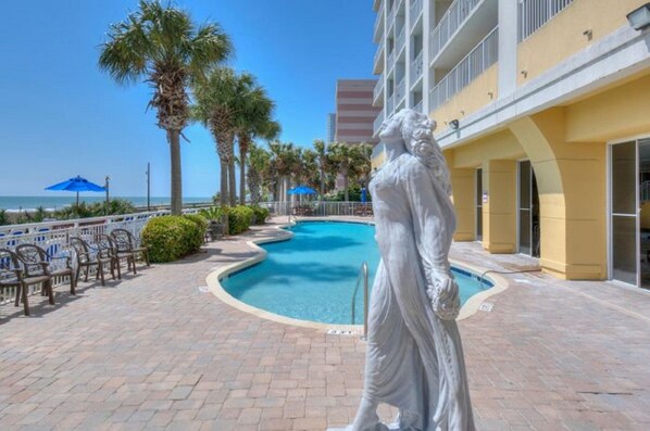 Relax by the serene pool surrounded by palm trees and a statue, with comfortable seating and umbrellas nearby.