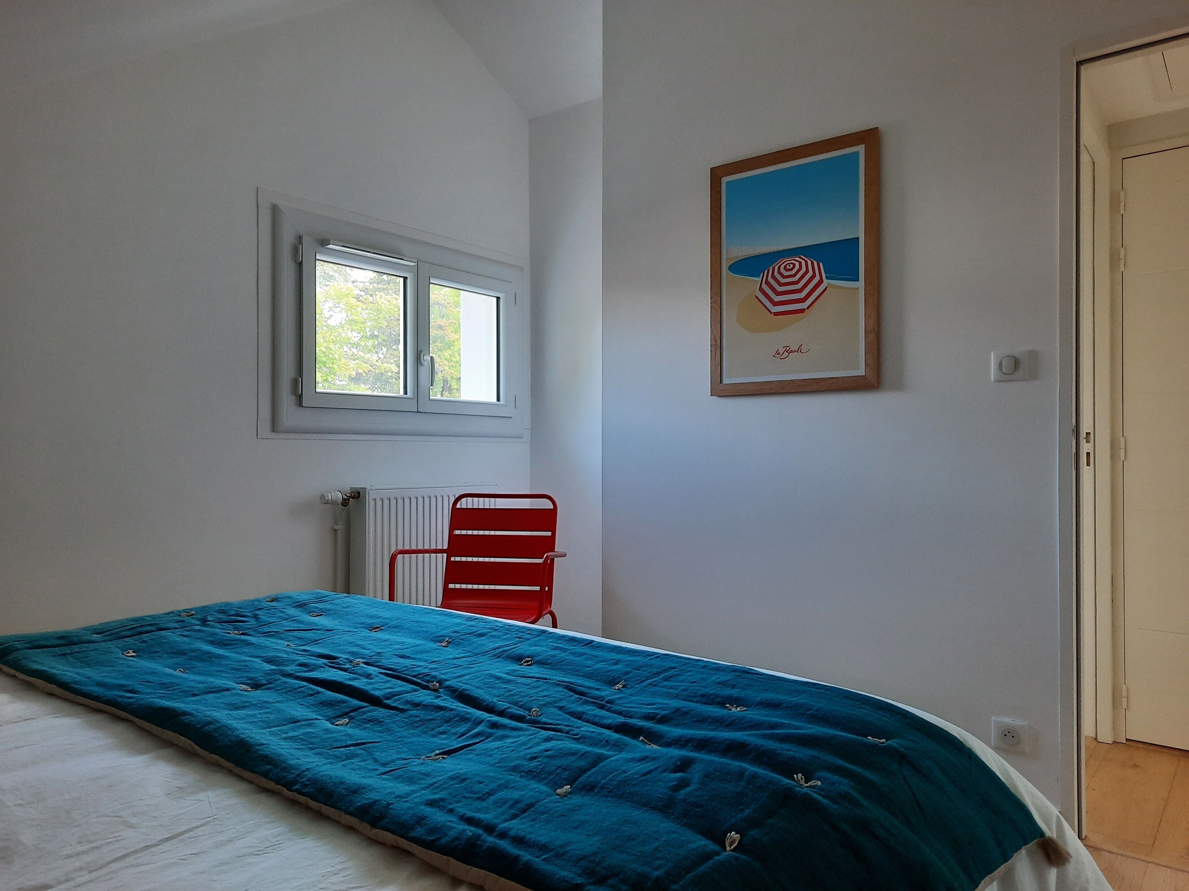 Quarto com cama, janela, radiador, uma cadeira vermelha e um quadro na parede.
