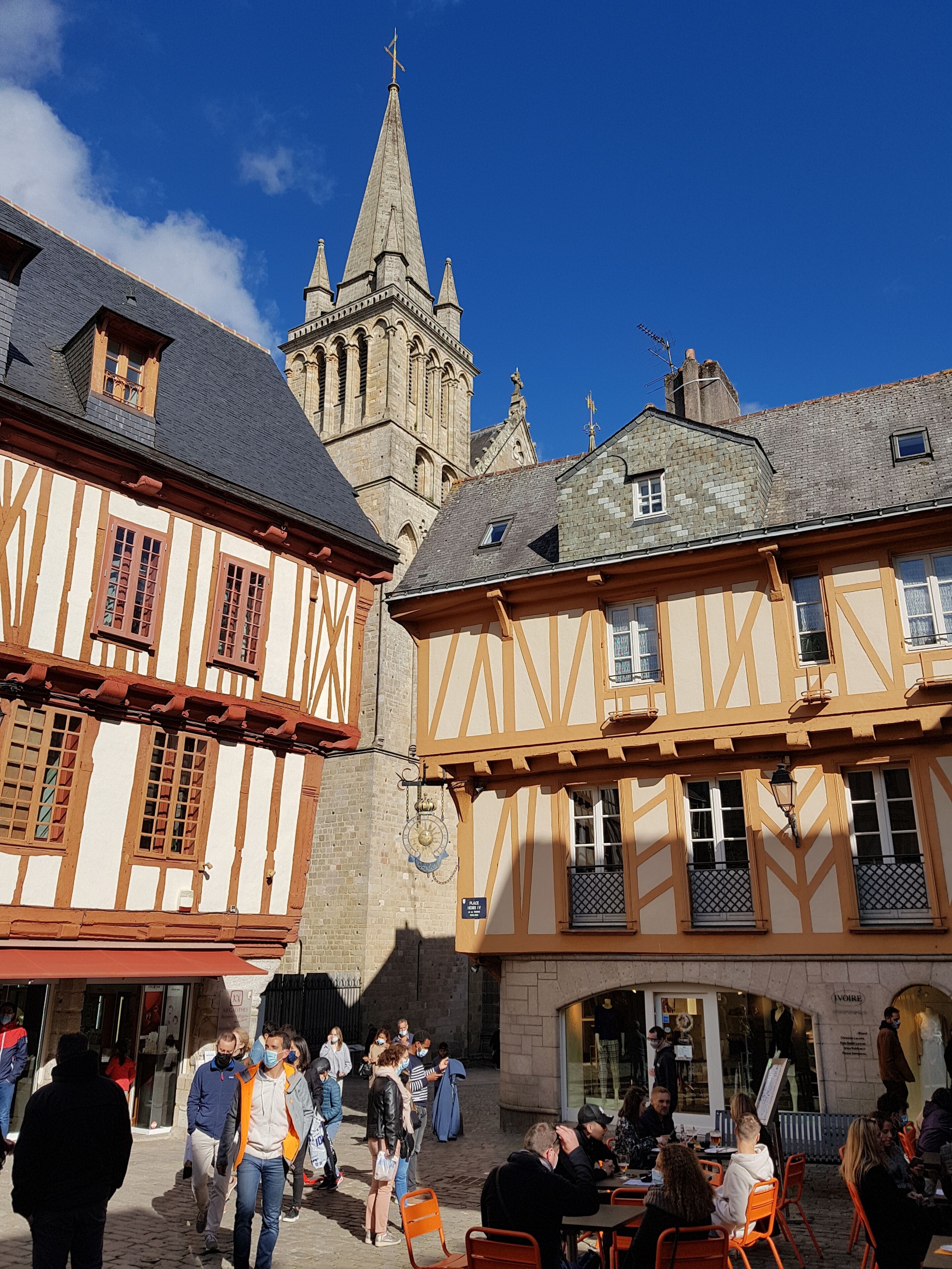 Una calle europea tradicional con edificios de entramado de madera, una torre de iglesia y gente caminando y cenando al aire libre.