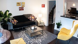 Un salon moderne avec un canapé noir, une table basse en verre et un fauteuil jaune. Un escalier mène à la cuisine.