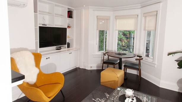 Un salon moderne avec un téléviseur à écran plat, un fauteuil jaune, une table à manger ronde et une fenêtre donnant sur un paysage verdoyant.