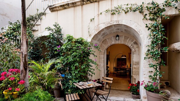 An outdoor patio with a wooden table and chairs, surrounded by potted plants and flowers. - CharmSuites - charming flat in Ortigia (Siracusa)