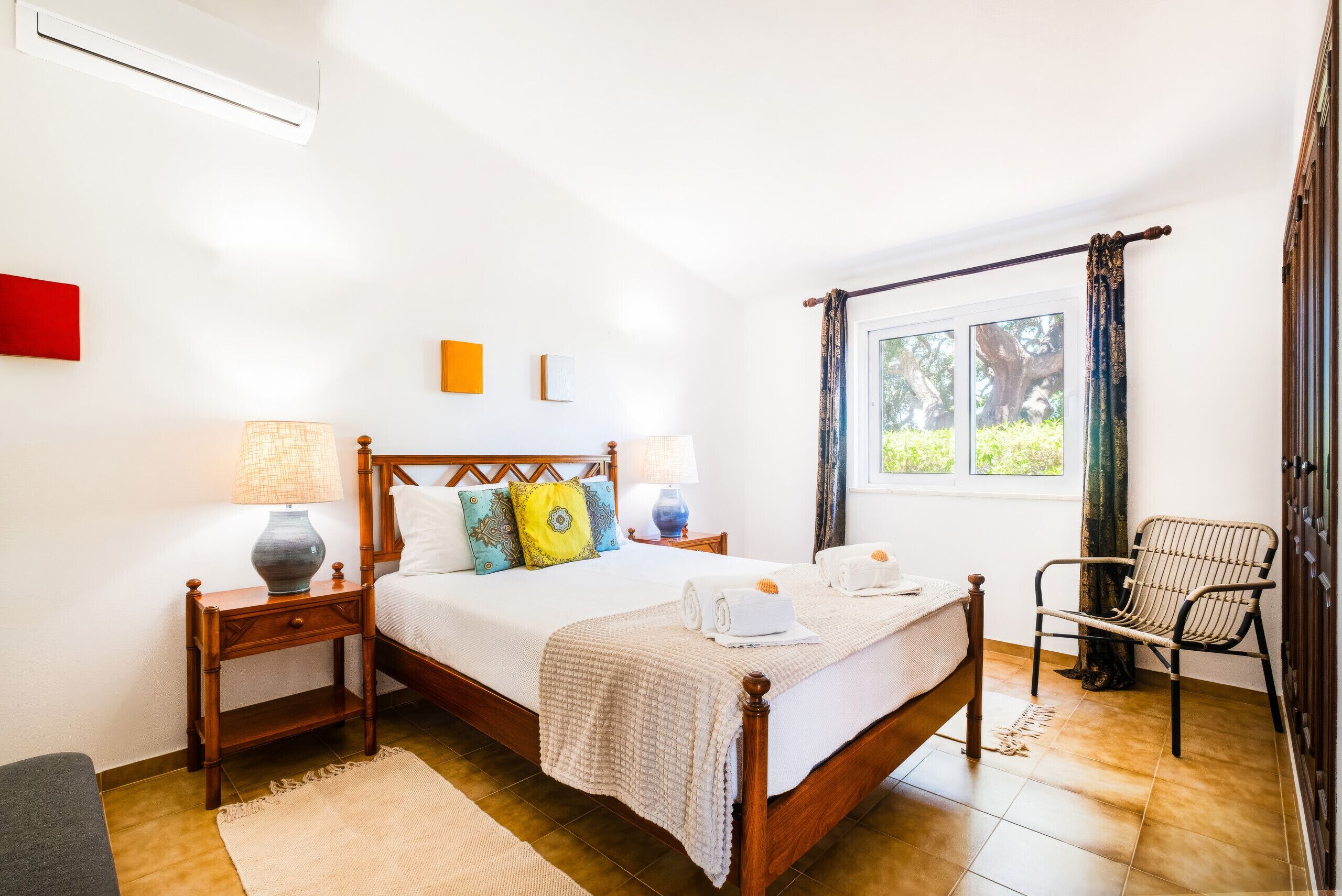 A well-lit bedroom with a wooden bed, two bedside tables, a chair, and a window with curtains.