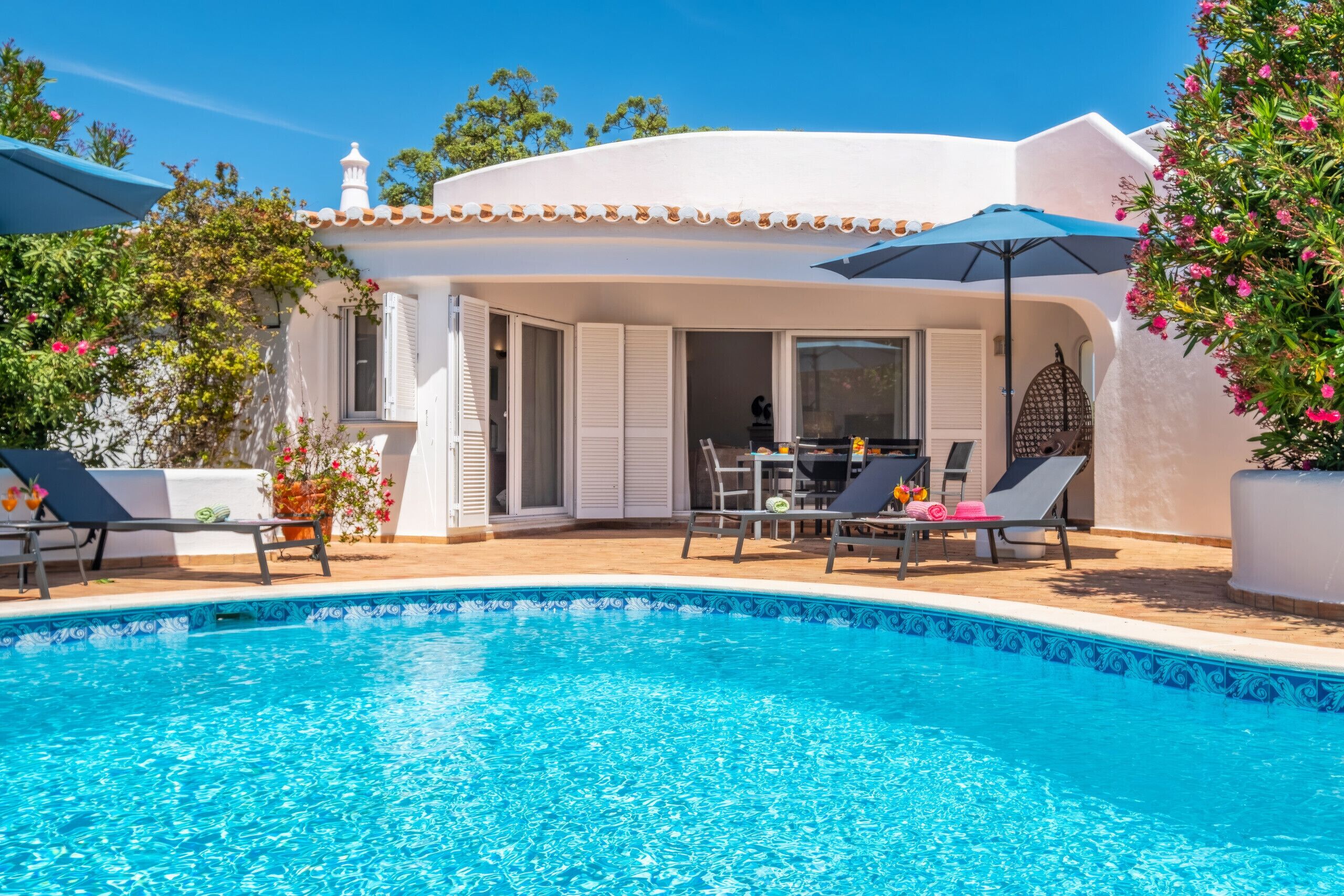 A poolside area with a clear blue pool, sun loungers, and a covered seating area with a table and chairs.