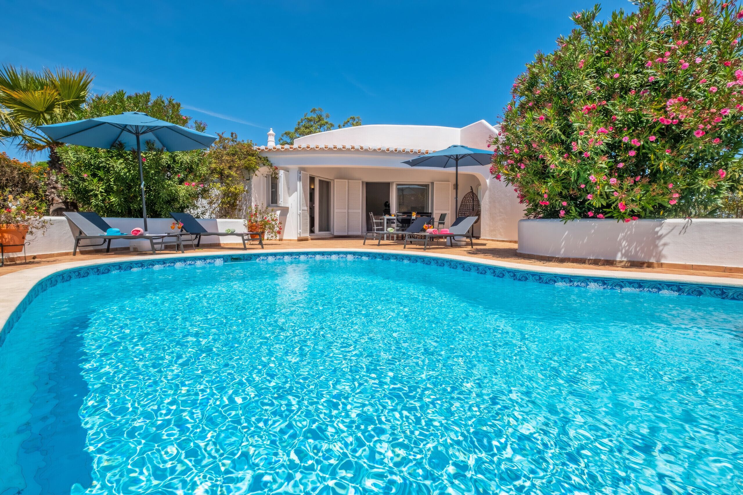A swimming pool with lounge chairs and umbrellas, surrounded by greenery and a building with a tiled roof.
