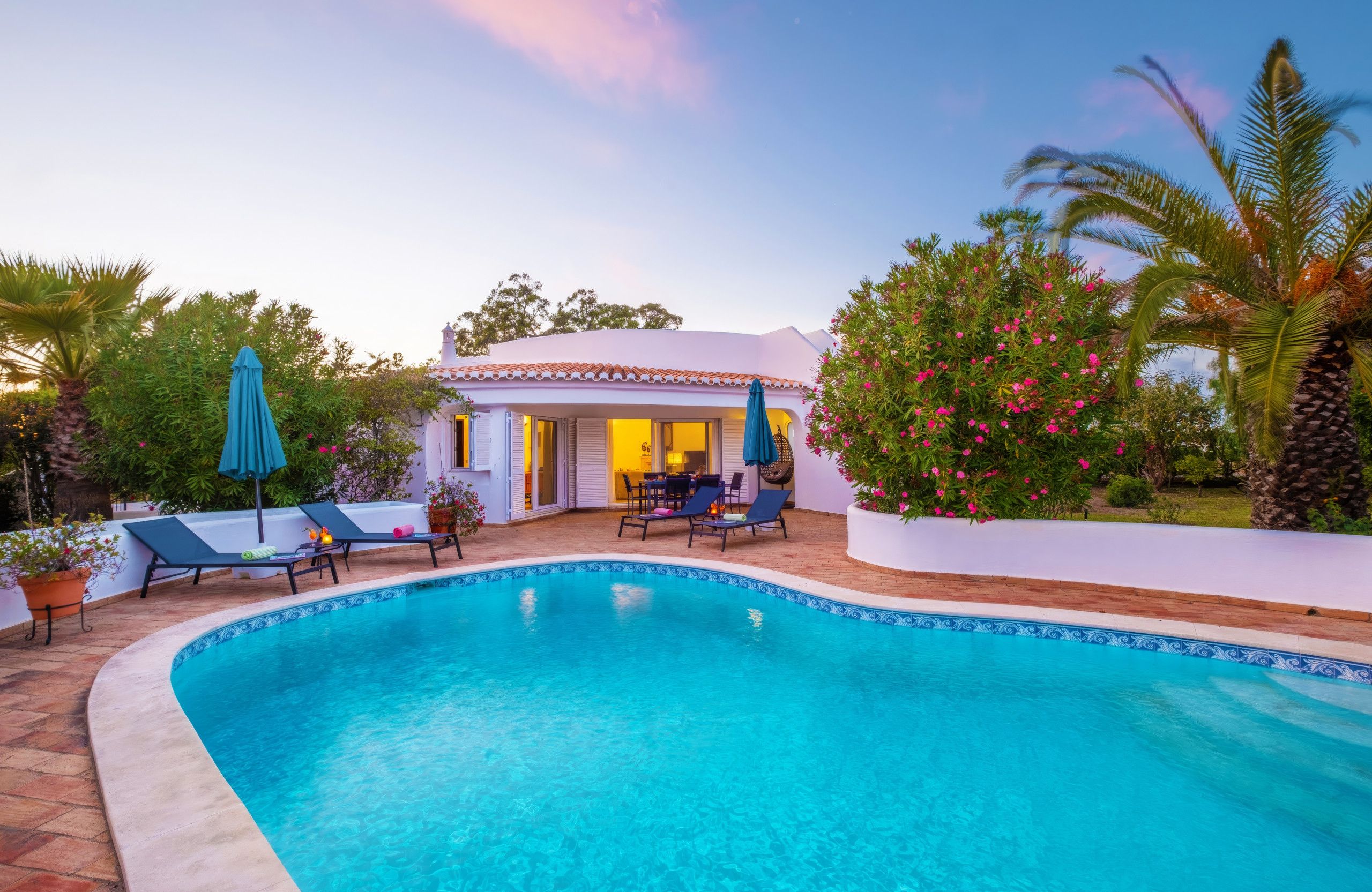 A swimming pool with lounge chairs, a covered seating area, and a white building with a tiled roof.