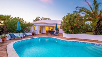 A swimming pool with lounge chairs, a covered seating area, and a white building with a tiled roof.