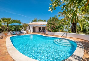 A clear blue swimming pool with lounge chairs and umbrellas, surrounded by lush greenery and a white building with a tiled roof.