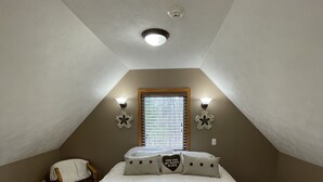A bedroom with a sloped ceiling, a bed with white bedding, two wall-mounted light fixtures, a window with blinds, and a small chair. - Perfect Getaway! Secluded Modern 3 Bedroom, 2 Bath Cabin on 12.5 Private Acres (Baldwin)