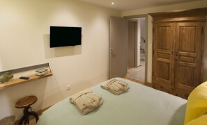 A bedroom with a bed, a TV mounted on the wall, a wooden wardrobe, and a shelf with a remote control.