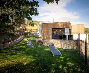 Een modern overdekt terras met twee lounge fauteuils, een stenen muur en een houten gebouw met een groot raam.