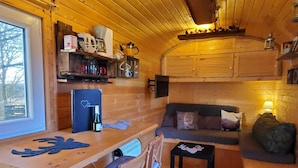 A cozy cabin interior with a wooden interior, a dining area with a table and chairs, a sofa, and a small kitchen area with shelves and appliances. - Gemütlicher Schäferwagen in der Eifel (Nettersheim)